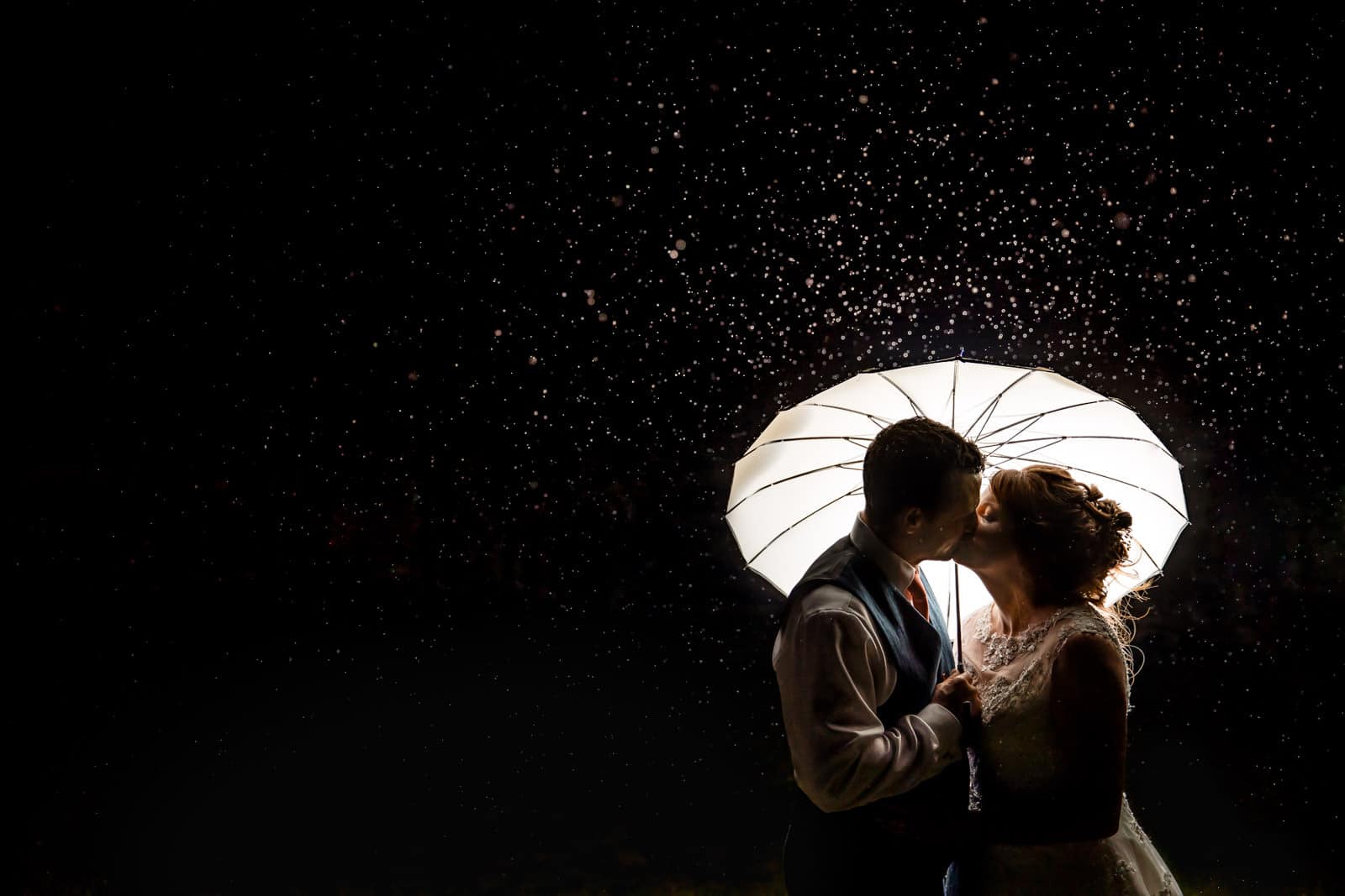 A bride and groom share a tender kiss under an umbrella in the rain beautifully captured by the Dodford Manor Wedding Photographer.